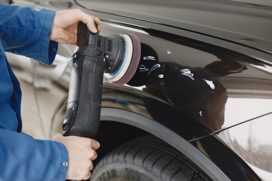 A person in protective gloves carefully sanding a car's body panel to remove loose paint and prepare the surface for primer.