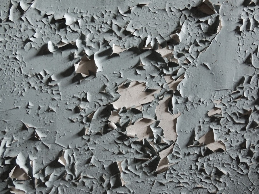 Person using a scraper tool to remove loose, peeling paint from a wall surface.