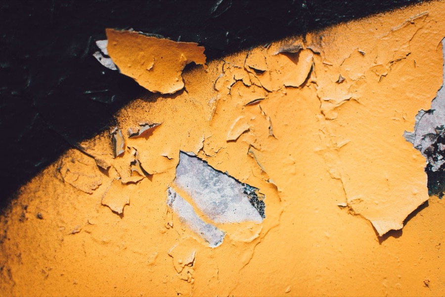 A person's hand using a paint scraper to carefully remove loose, peeling paint from a wall.