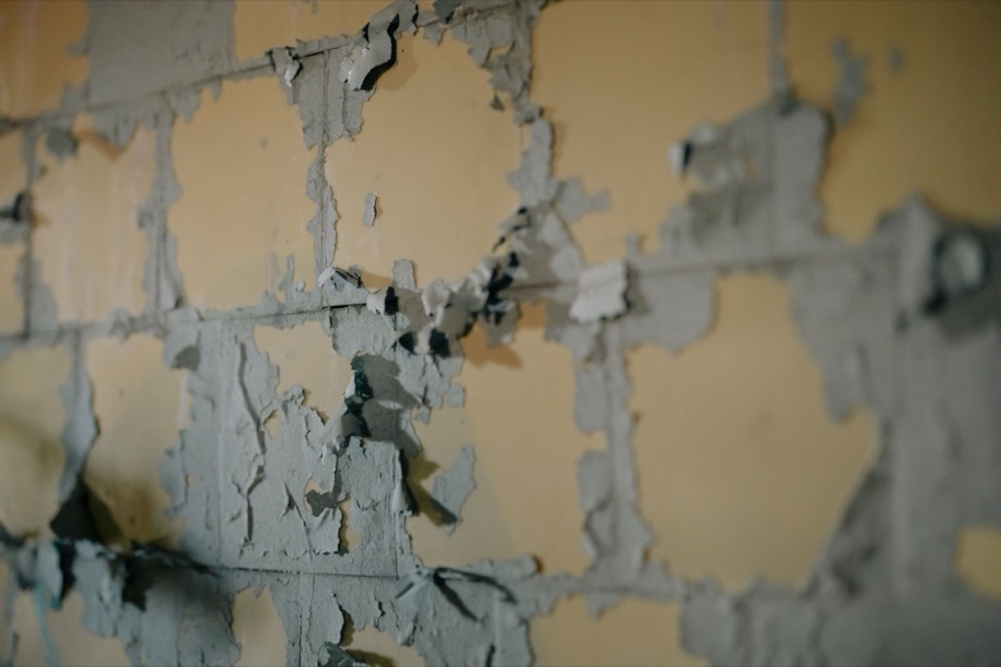 A person's hand using a paint scraper to remove loose, peeling paint from a wall surface.